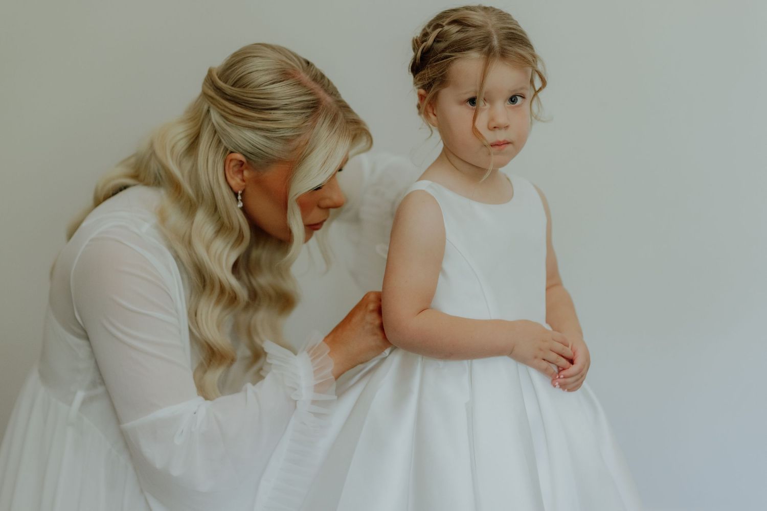 Bride in white gown adjusting the dress of a young flower girl against a light background.