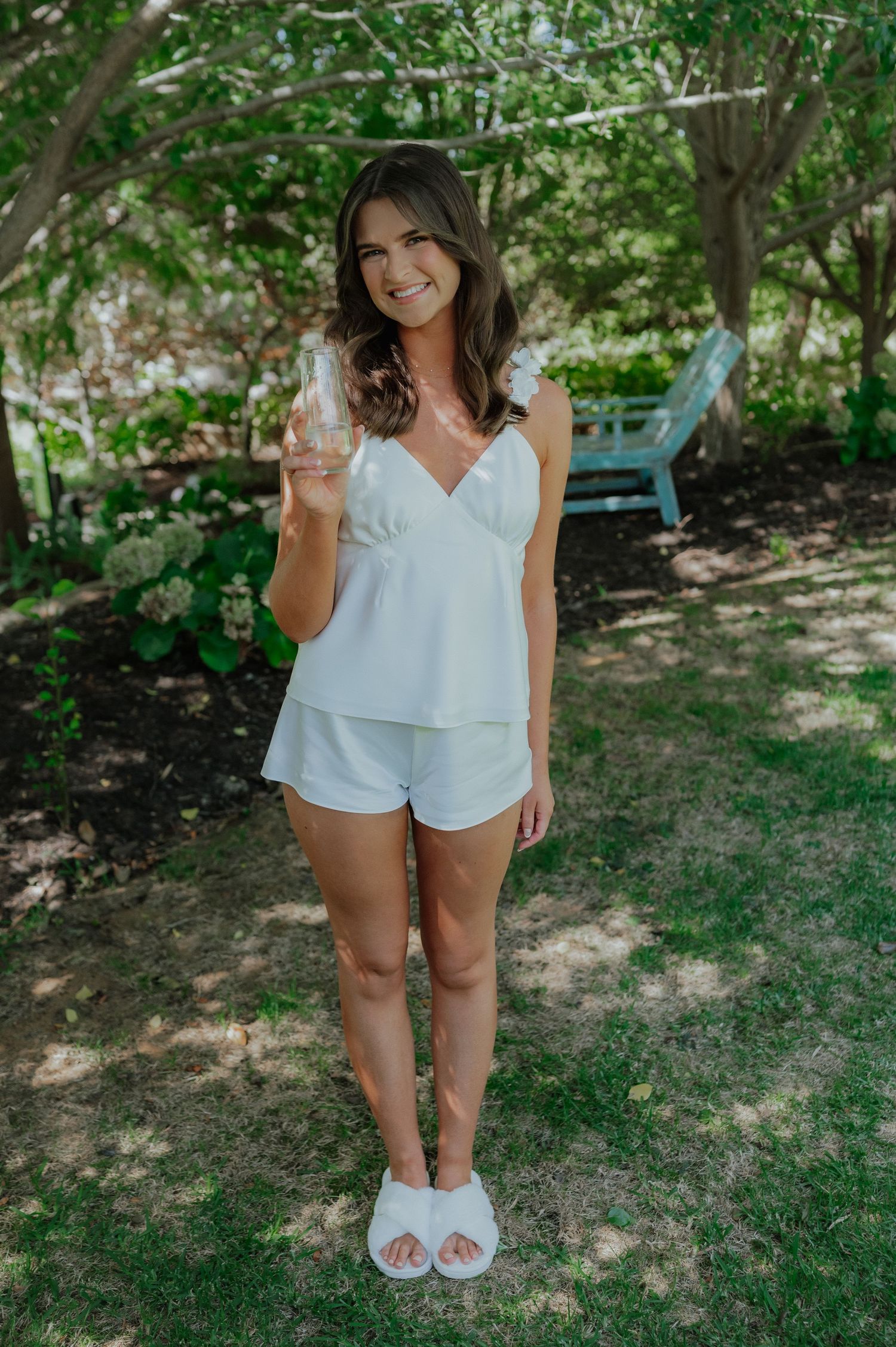 Smiling bride in white loungewear holds champagne glass while standing in lush garden before wedding.