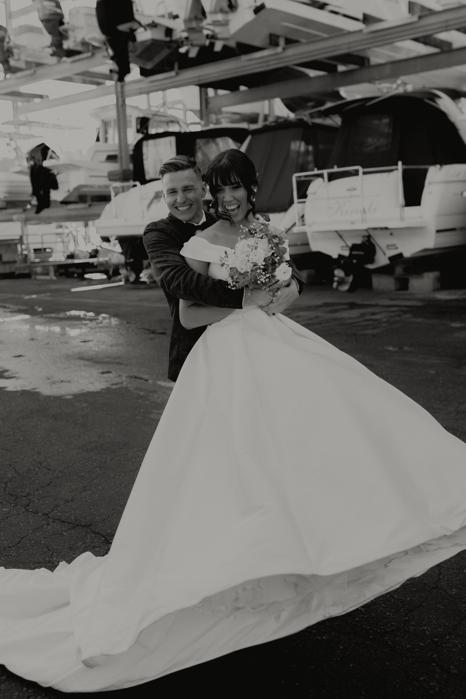 Black and white photo of smiling bride and groom embracing outdoors, brides gown with long flowing train.