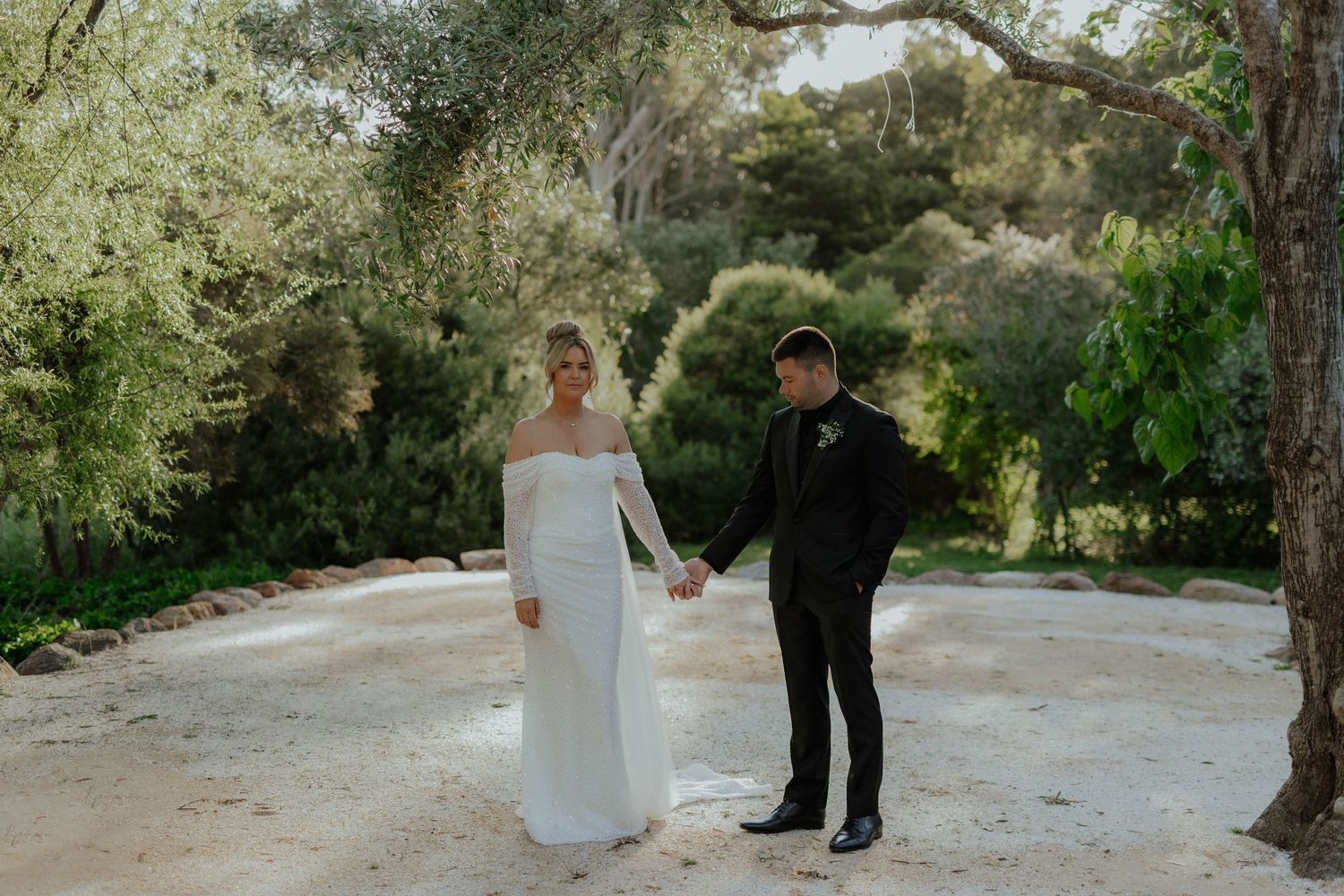 Bride in long-sleeve white gown and groom in black suit hold hands in lush garden.