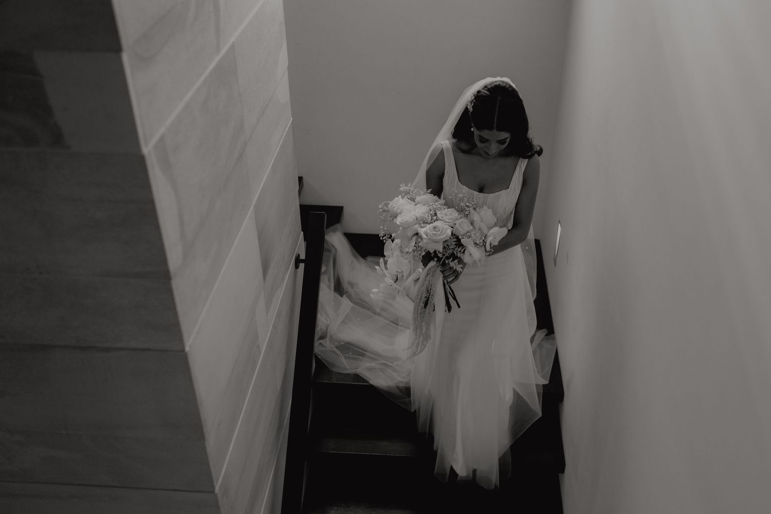 Black and white photo of bride in wedding gown descending staircase while holding white bridal bouquet.