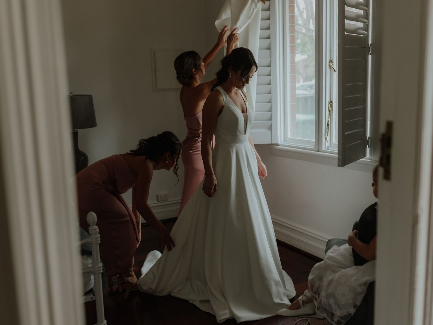 Bridesmaids helping bride into her white wedding gown in a bright room with shuttered windows.