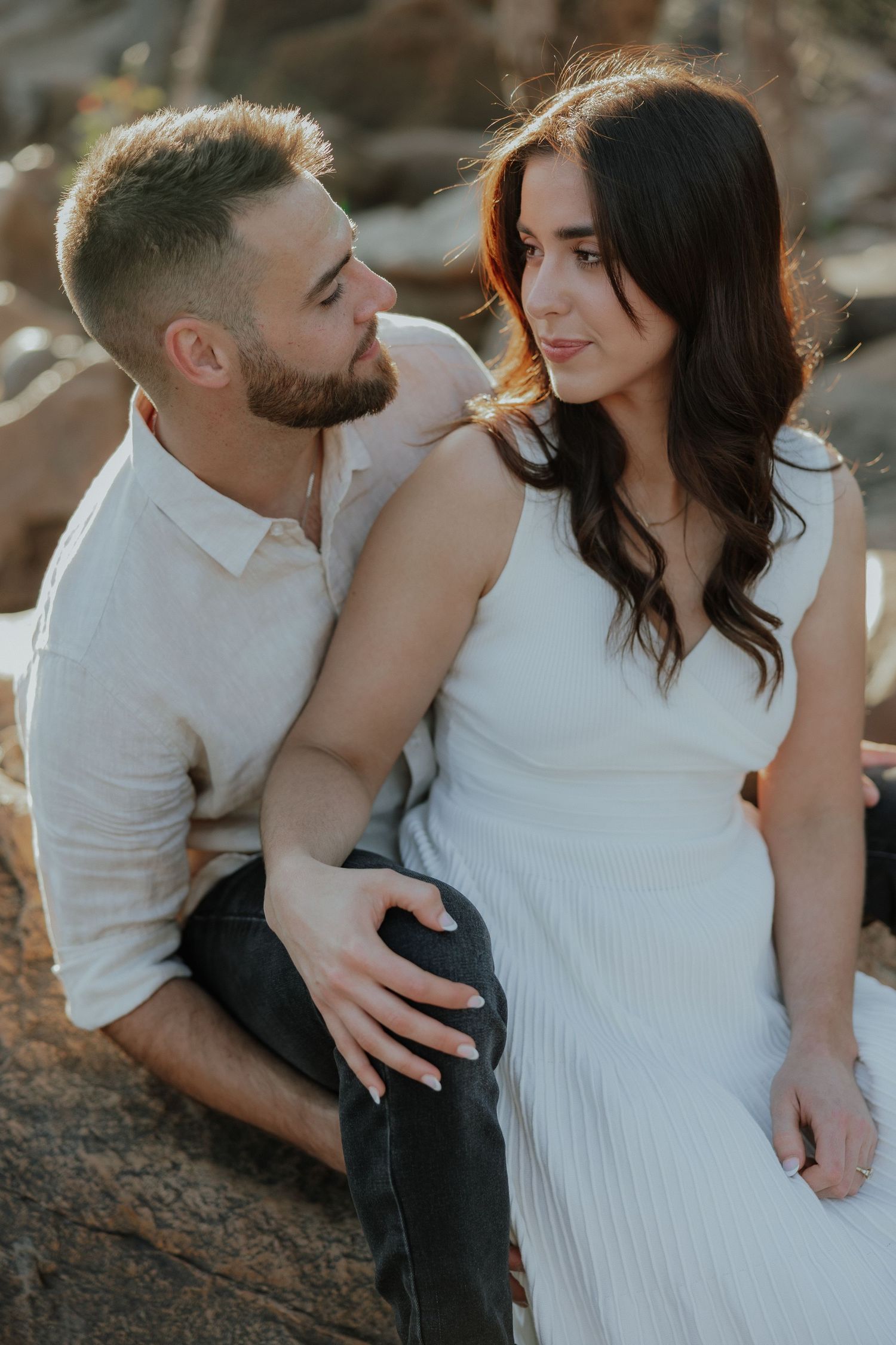 Man with beard tenderly embraces woman in white dress while sitting on sun-warmed rocks outdoors.