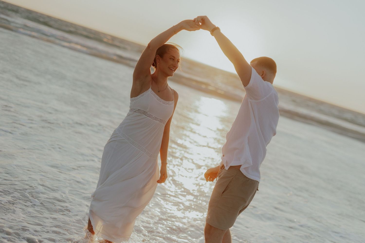 Couple dancing on beach at sunset in white outfits, holding hands and forming an arch.