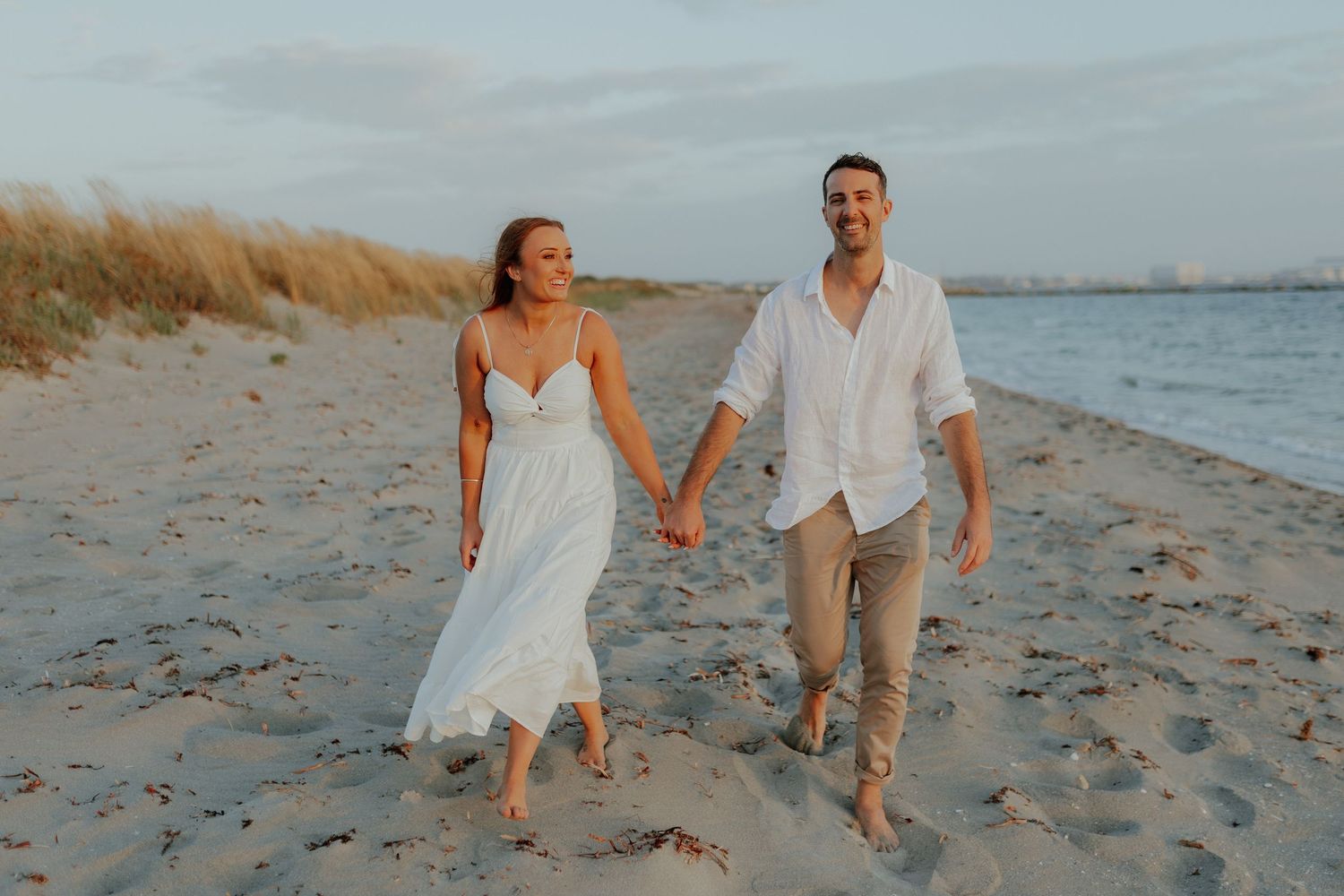 Smiling couple in white holding hands walking barefoot on sunny sandy beach.