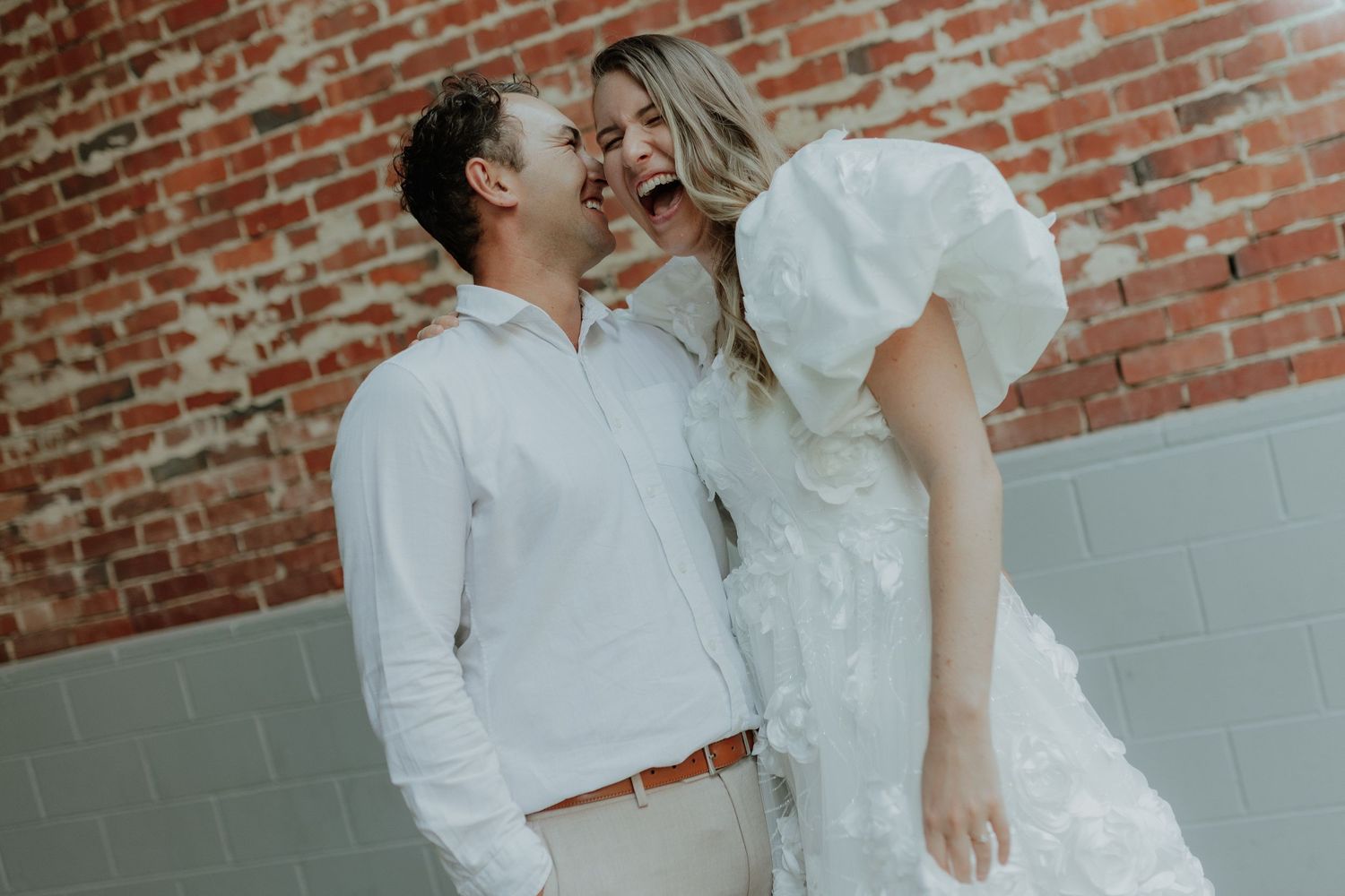 Laughing couple in white wedding attire posing playfully against a rustic red brick wall outdoors.