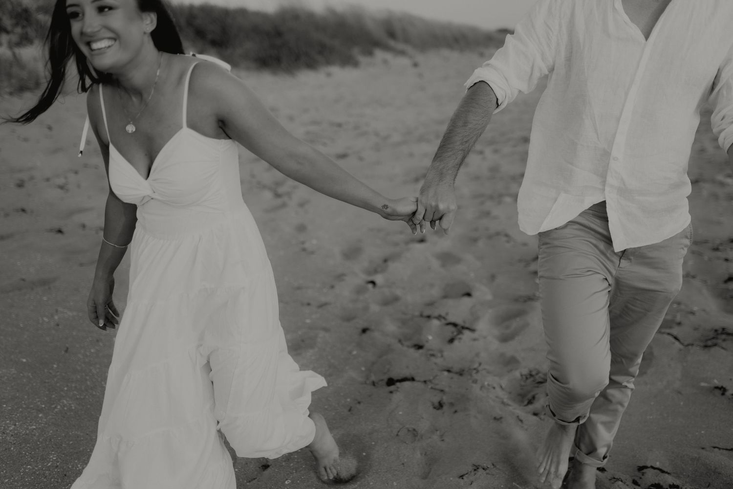Black and white photo of couple holding hands walking on sandy beach.
