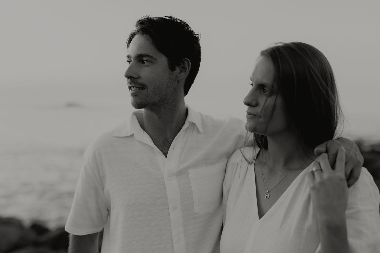 Black and white portrait of couple with man's arm around woman gazing toward ocean horizon.