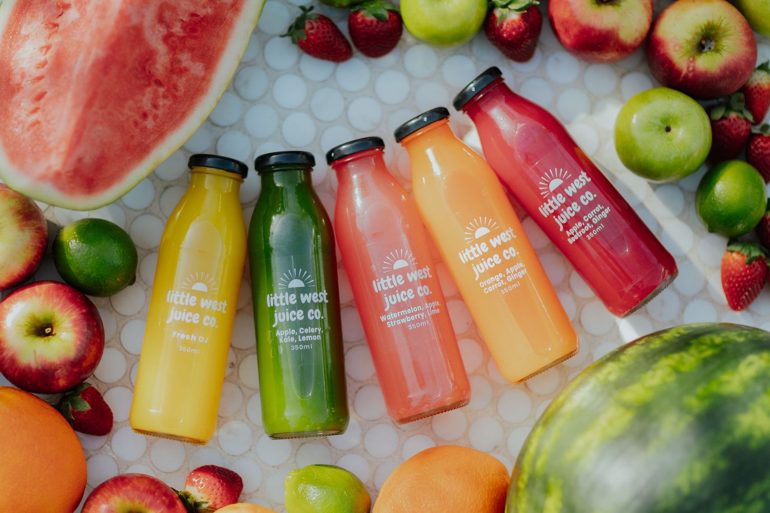 Five colorful Little West Juice Co. bottles surrounded by fresh watermelon, apples, and berries.