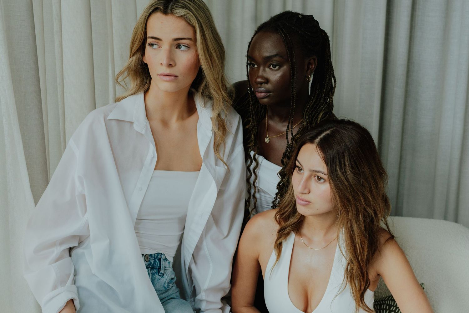 Three diverse women pose together in white outfits against a soft draped background.