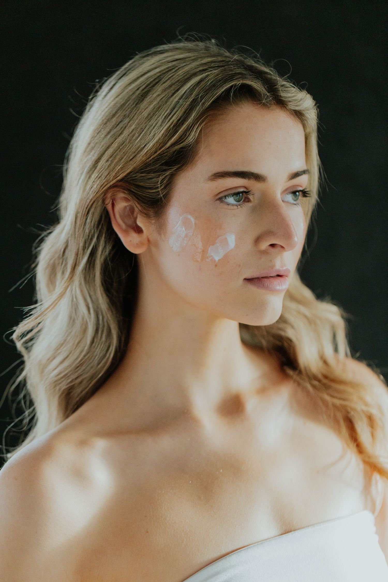 Blonde woman with white cream dots applied to her cheek for skincare routine against dark background.