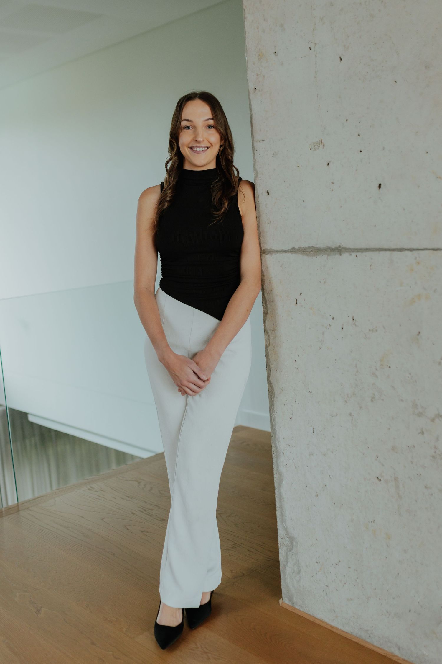 Professional woman in black top and white wide-leg pants posing in a modern office interior.