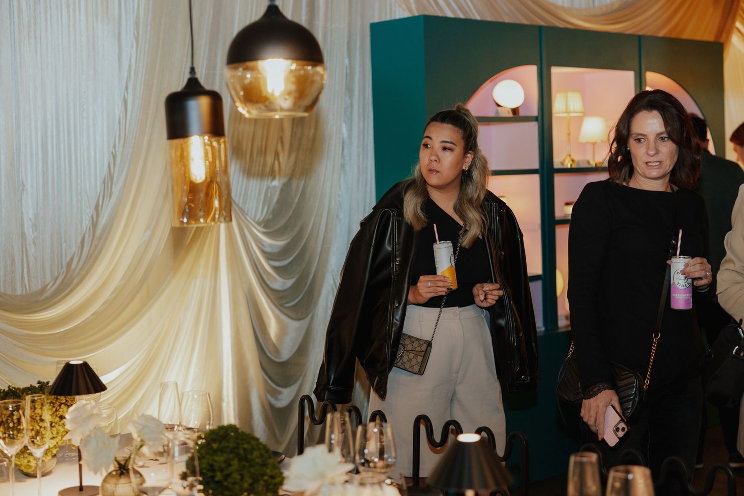 Two women in stylish outfits explore a decorated event space with pendant lights and table settings.