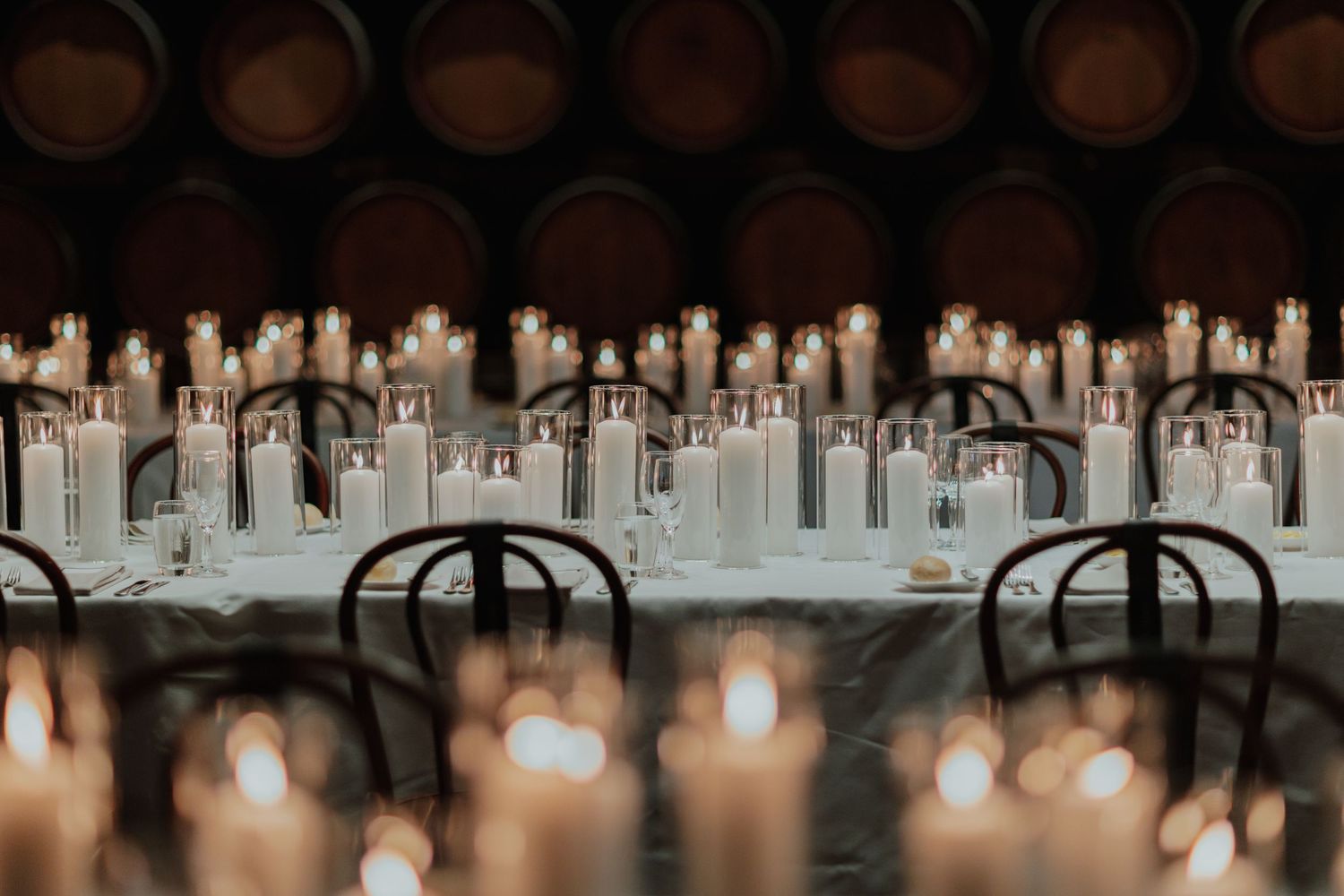 Elegant candlelit dinner table with rows of white pillar candles and wine barrels in background.