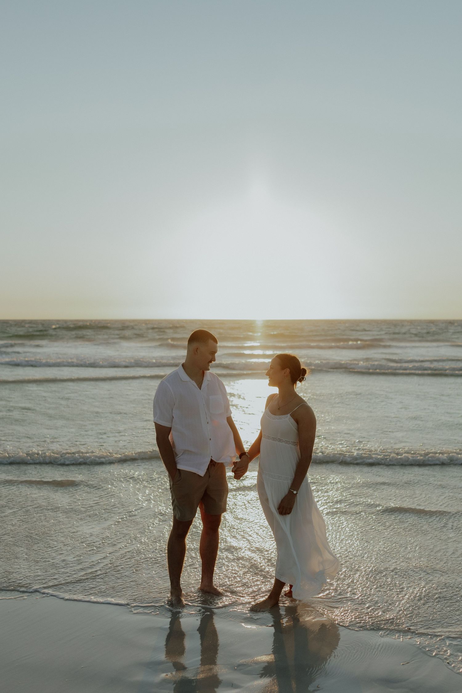 Couple in white holding hands on a beach at golden sunset with waves at their feet.