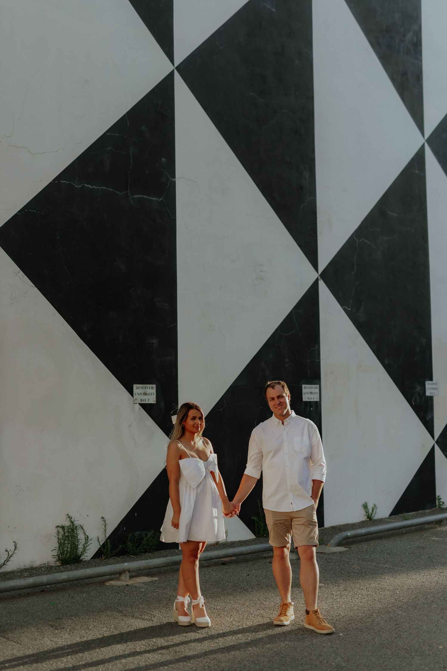 Couple holding hands in front of a bold black and white geometric mural on a sunny day.