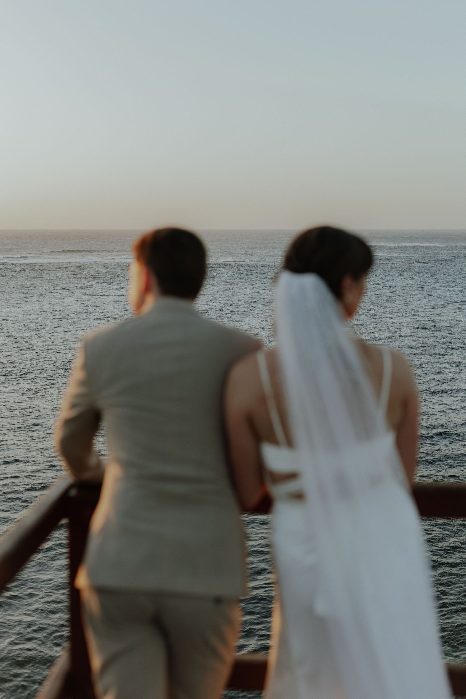 Bride in white gown with veil and groom in beige suit standing together overlooking calm ocean at dusk.