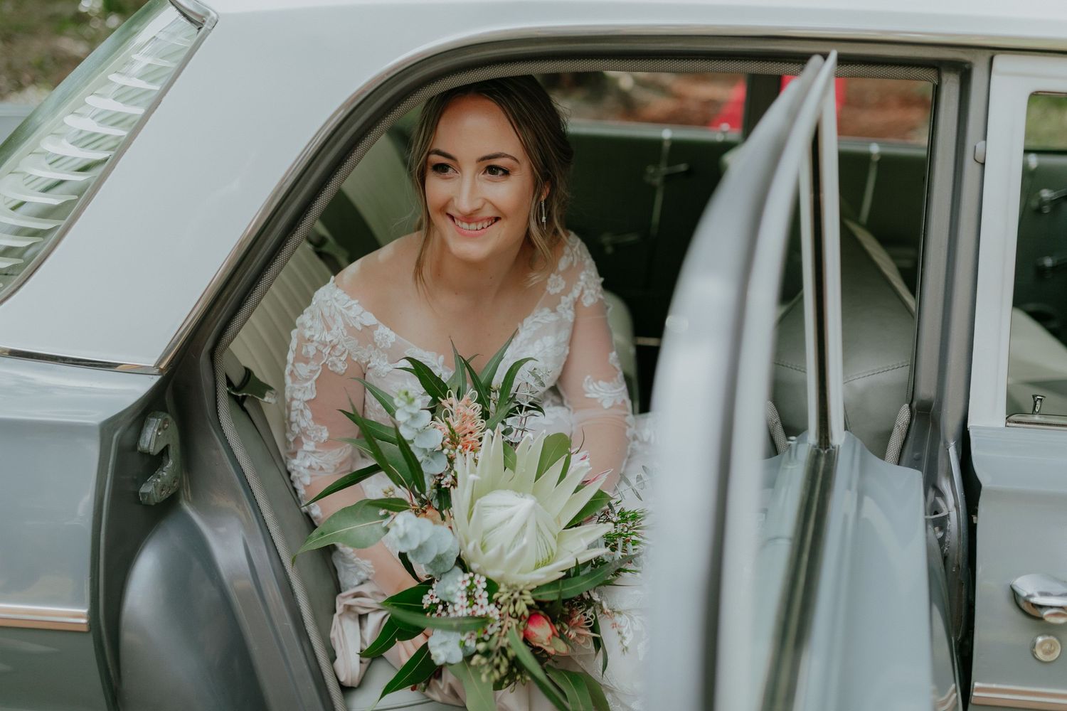 Radiant bride in lace gown holding tropical bouquet smiling from vintage silver wedding car.