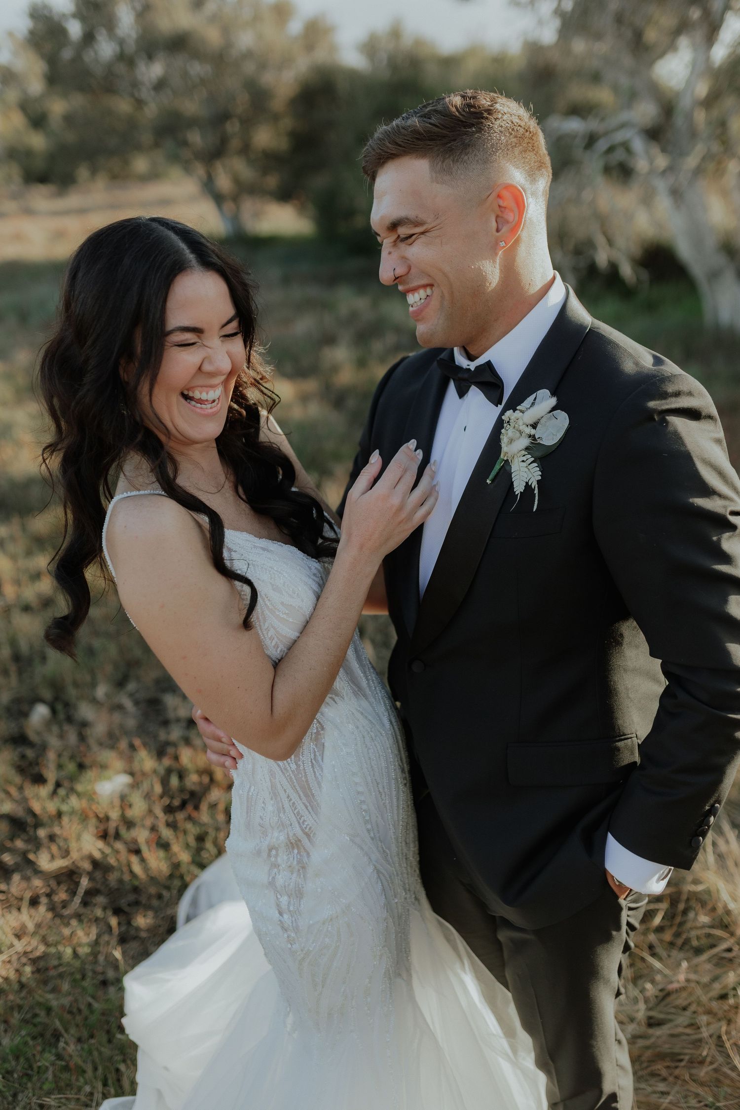 Bride and groom laughing together outdoors in wedding attire, black tuxedo and white lace gown.