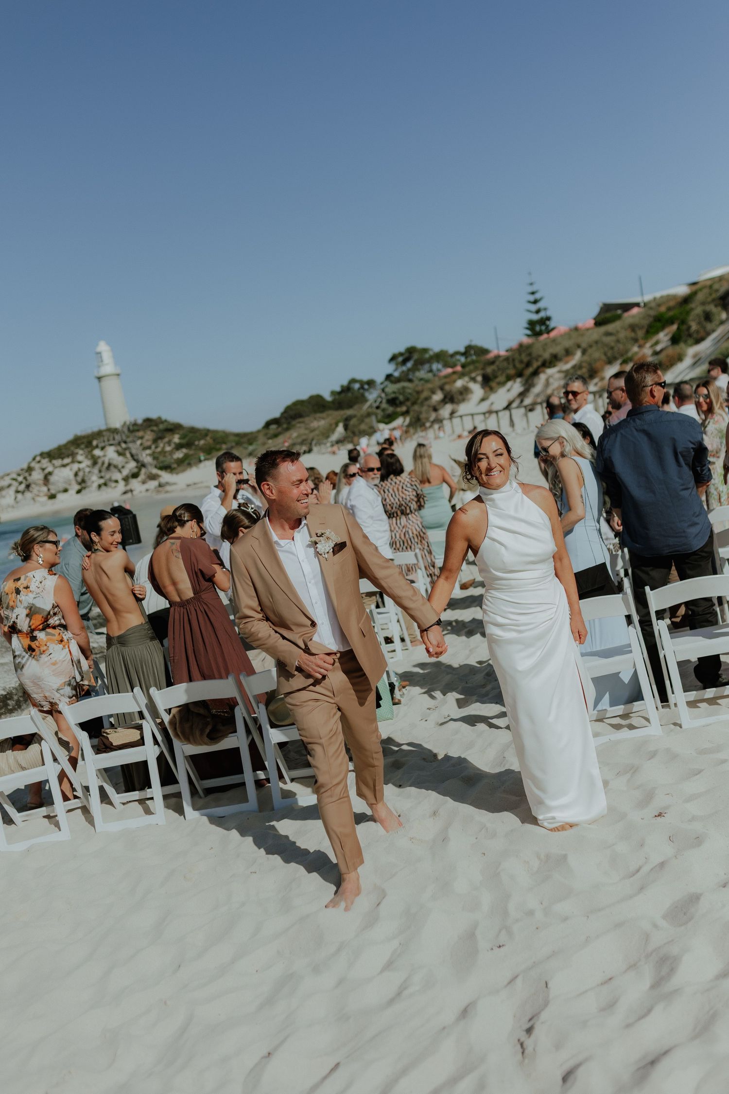 Joyful newlyweds walk down sandy beach aisle after wedding ceremony with lighthouse backdrop.