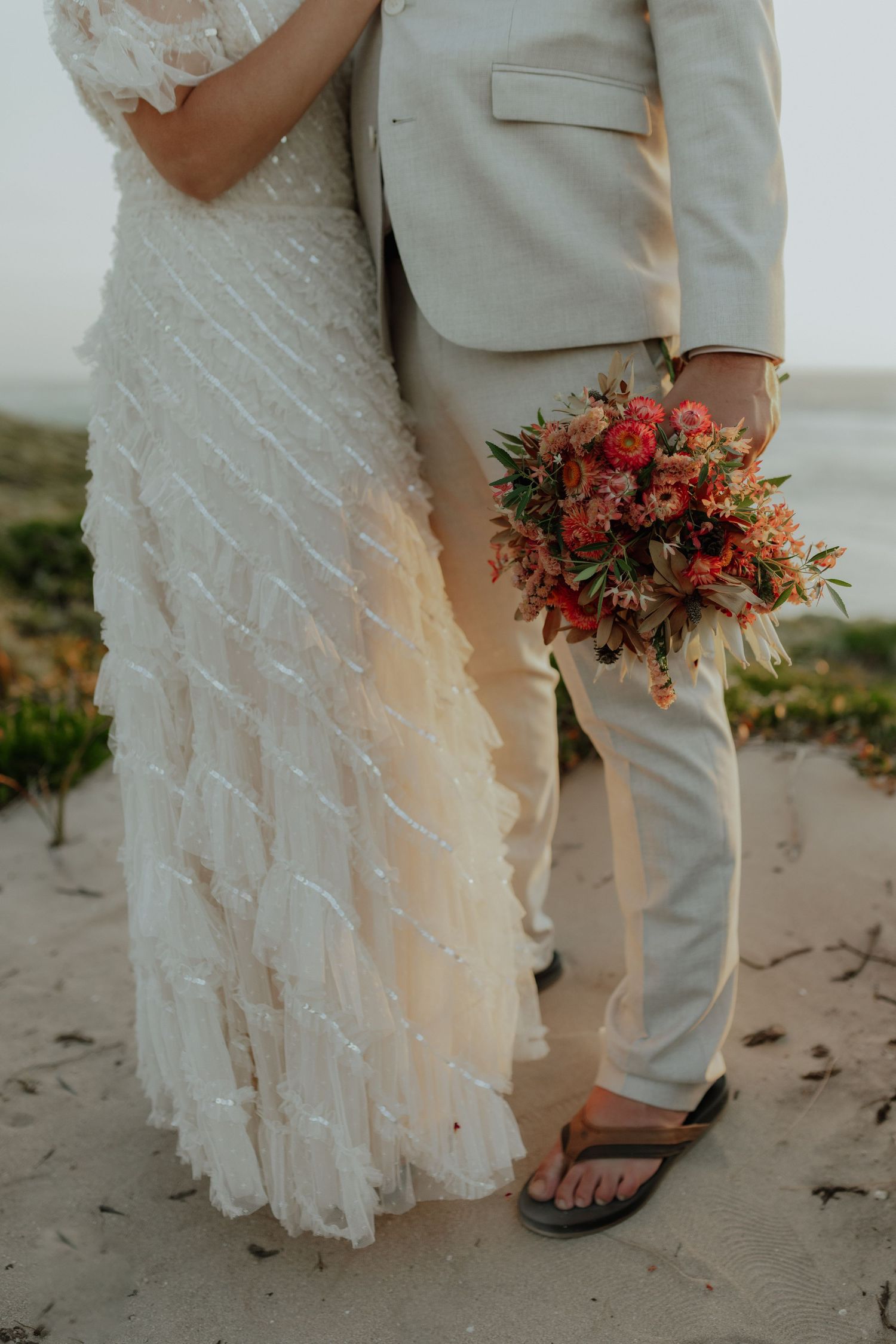 Bride and groom standing on sandy beach, groom holding vibrant red and orange bouquet.