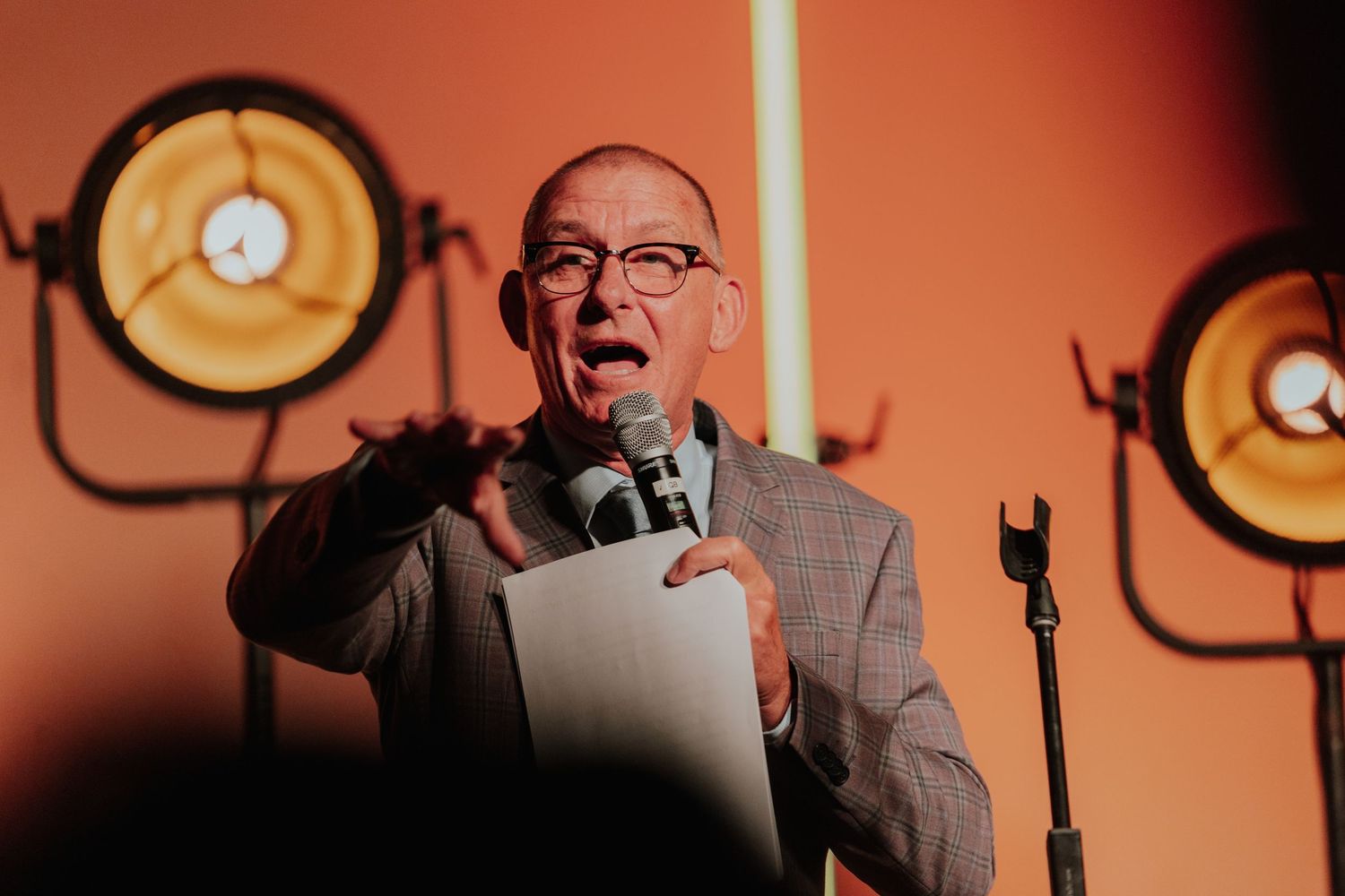 Bald man in grey suit speaking passionately into a microphone on stage with spotlights behind.