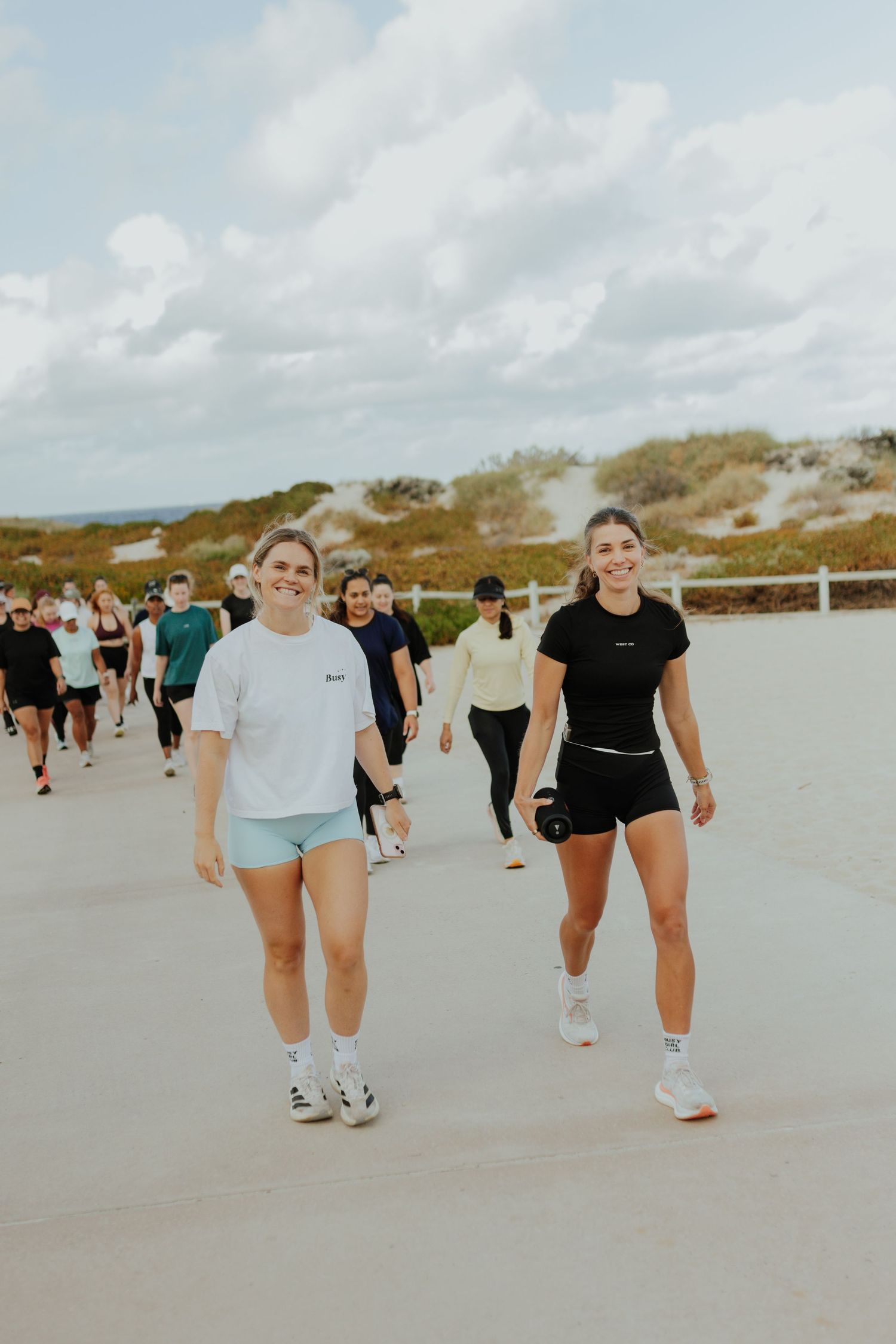 Two smiling women in athletic wear leading a group walk along a sandy beach path.