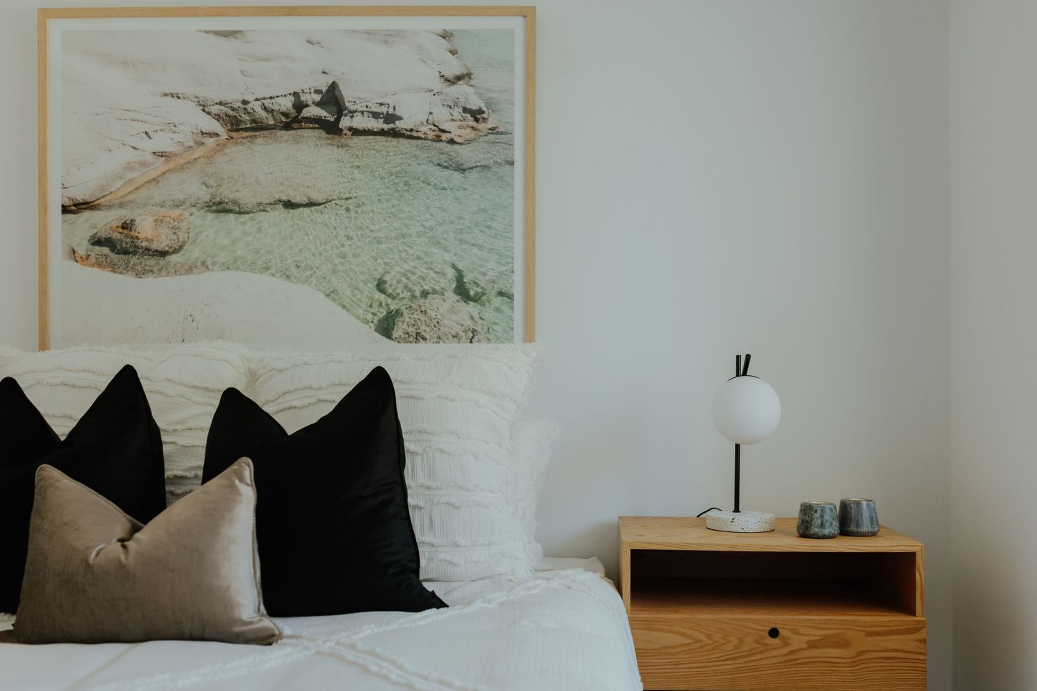 Minimalist bedroom with white bedding, black pillows, and a coastal landscape artwork above the bed.