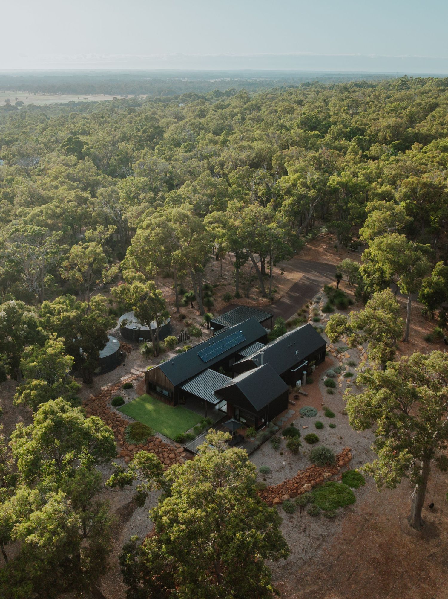 Aerial view of a modern black barn-style home nestled among dense Australian bushland with red dirt paths.
