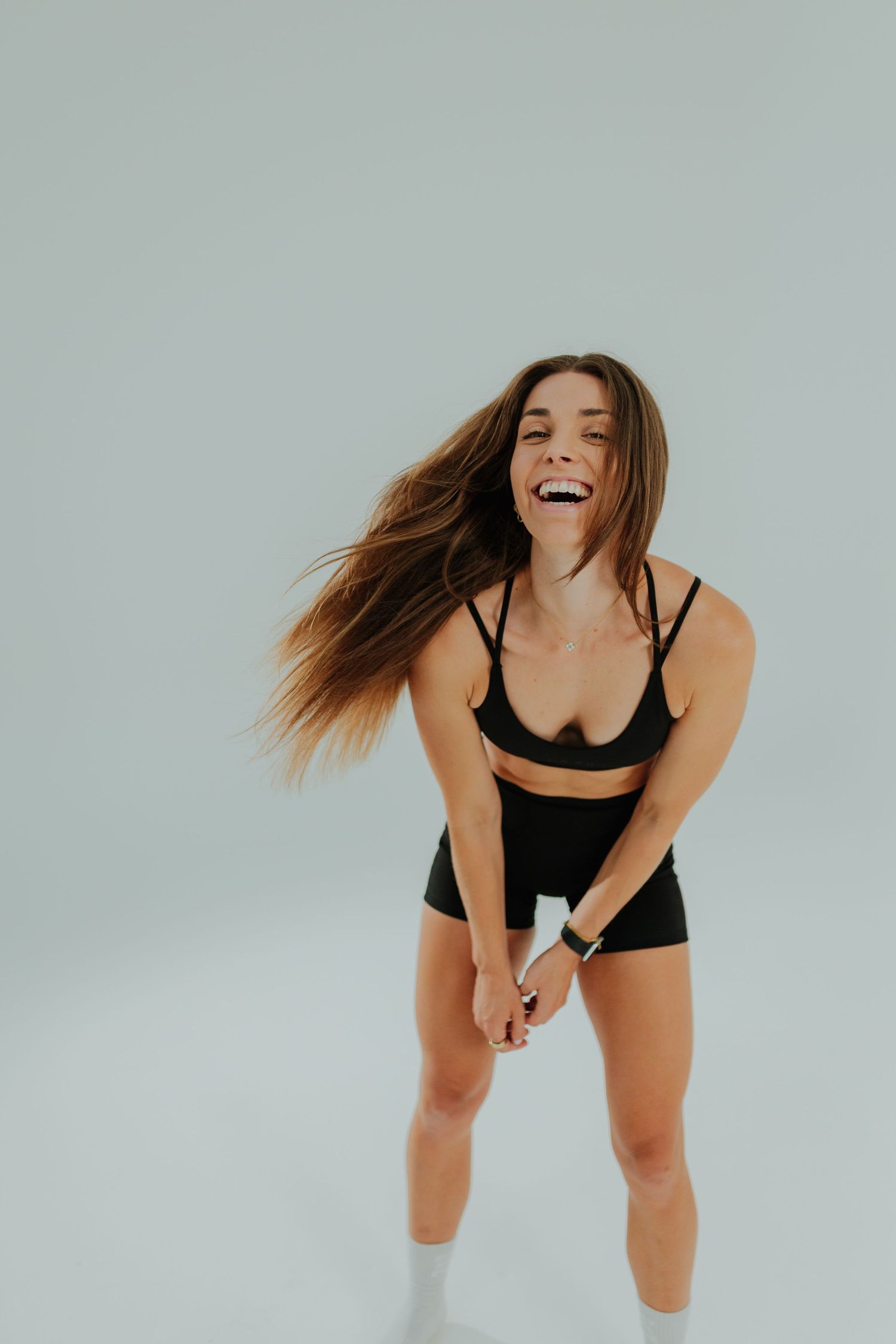 Athletic woman in black sports bra and shorts laughing with hair flowing against a light gray background.