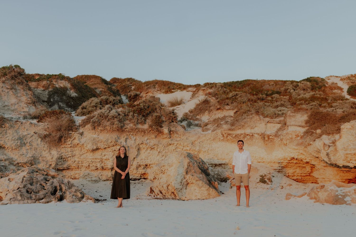 Couple standing apart on sandy beach at golden hour with colorful rocky cliffs in the background.