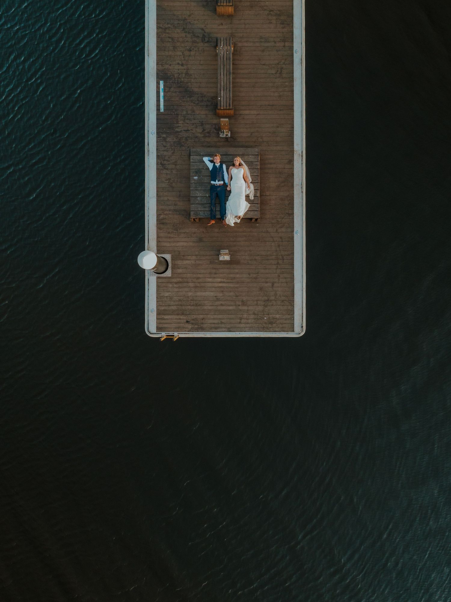 Aerial drone view of bride and groom lying on a wooden pier surrounded by dark water.
