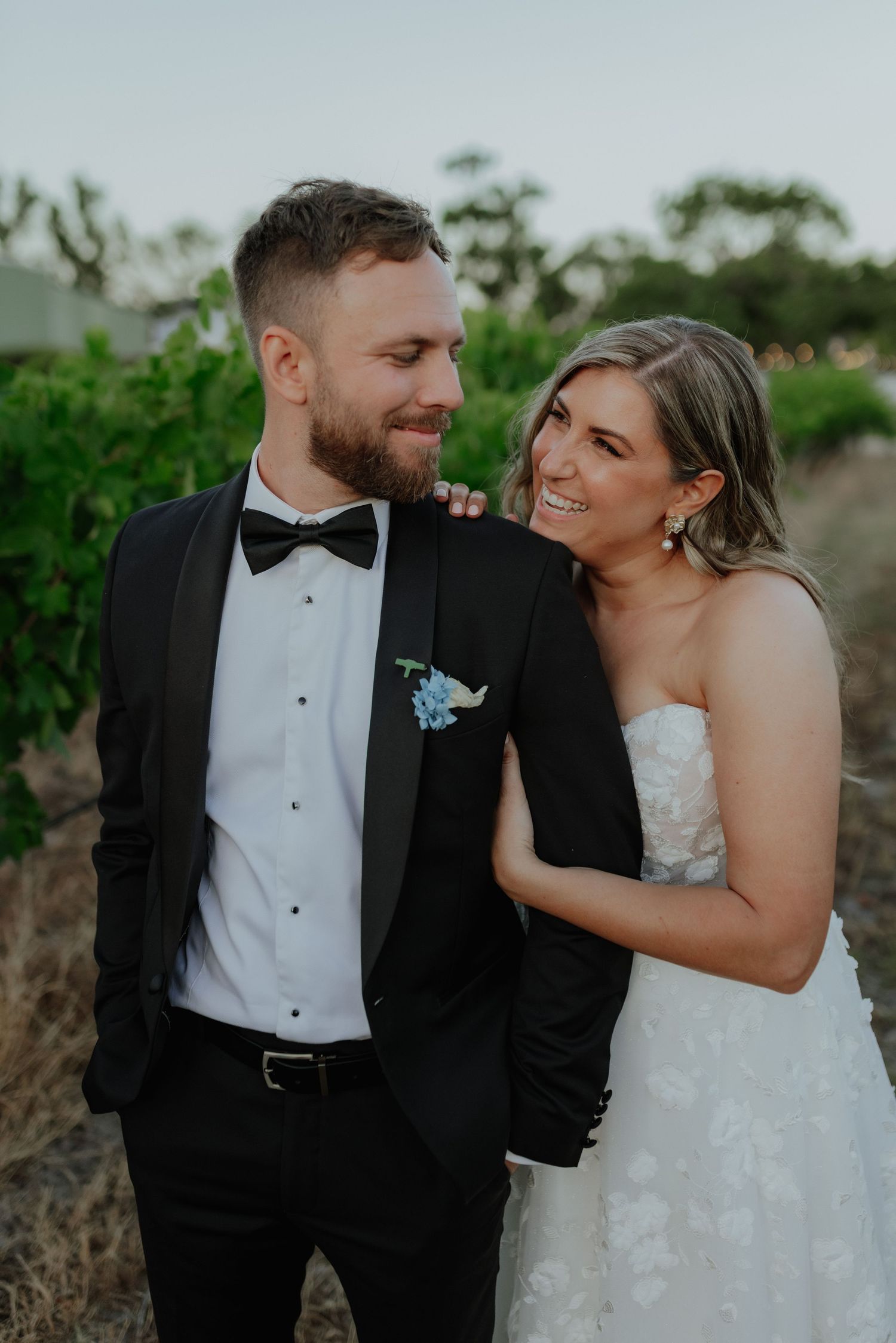 Smiling bride in strapless gown leans on groom in black tuxedo among vineyard rows.
