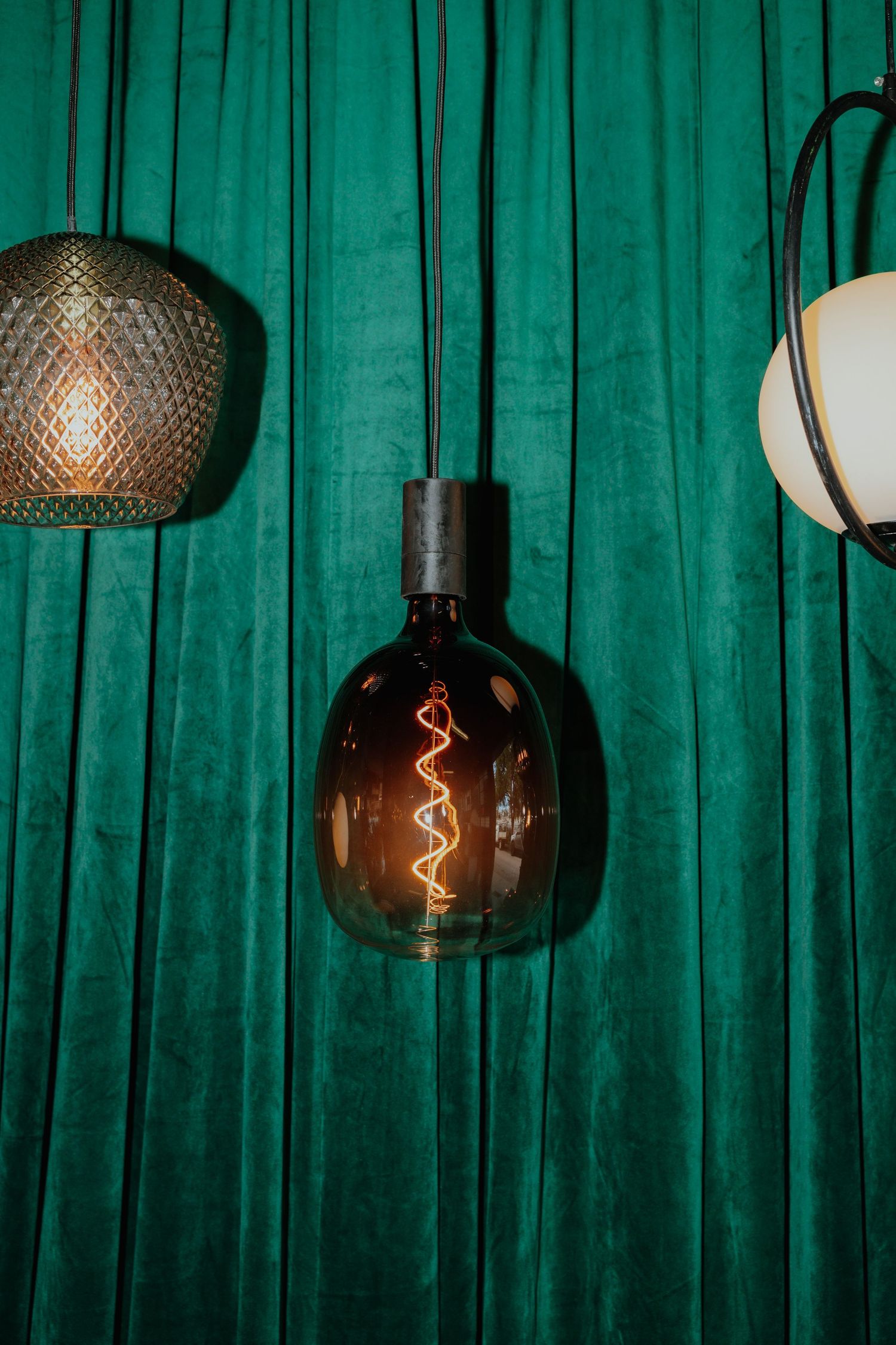 Vintage-style Edison bulb pendant light with spiral filament glowing against a deep green velvet curtain backdrop.