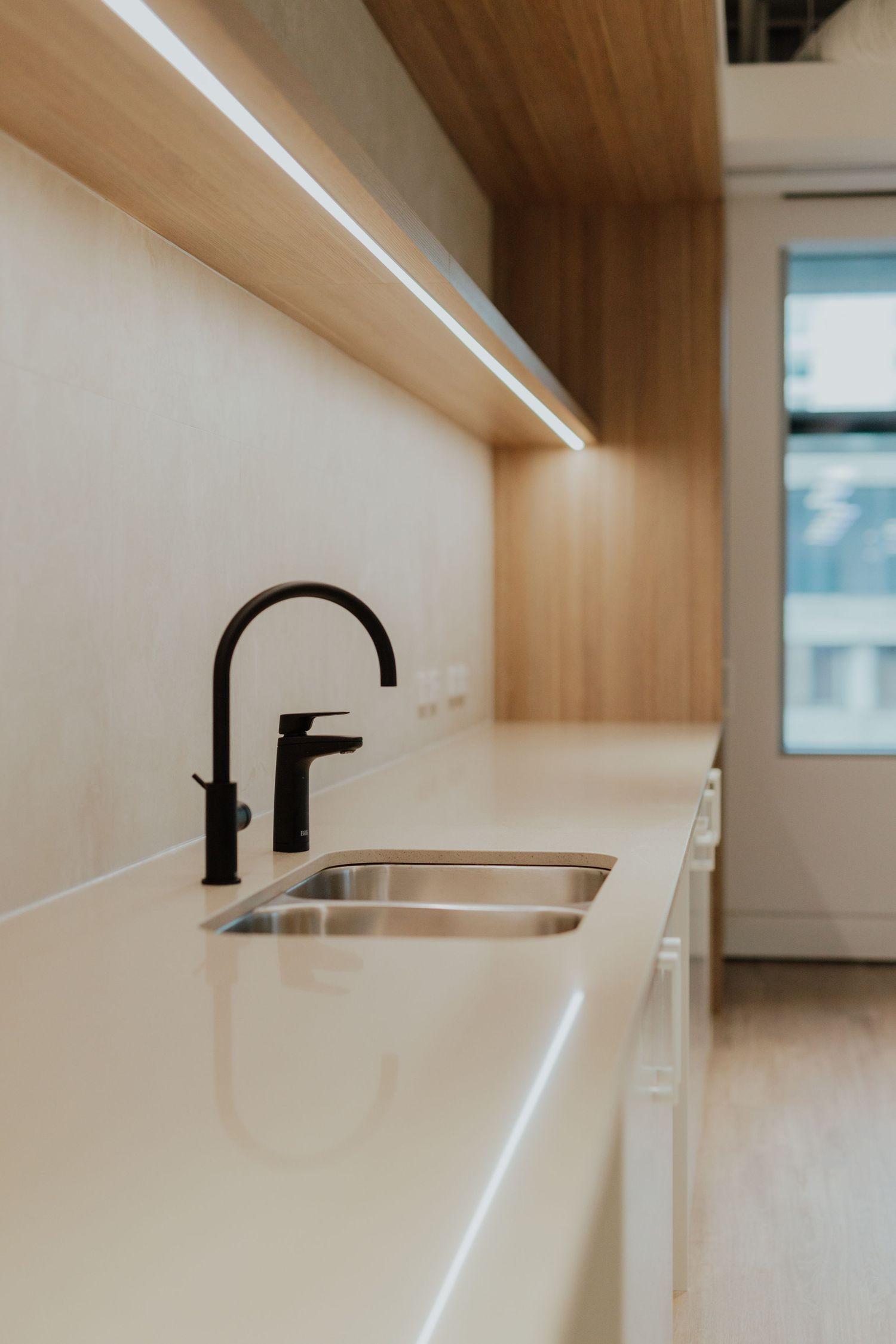 Minimalist kitchen butler pantry with white stone benchtop, stainless steel sink, black tap, and timber shelving.