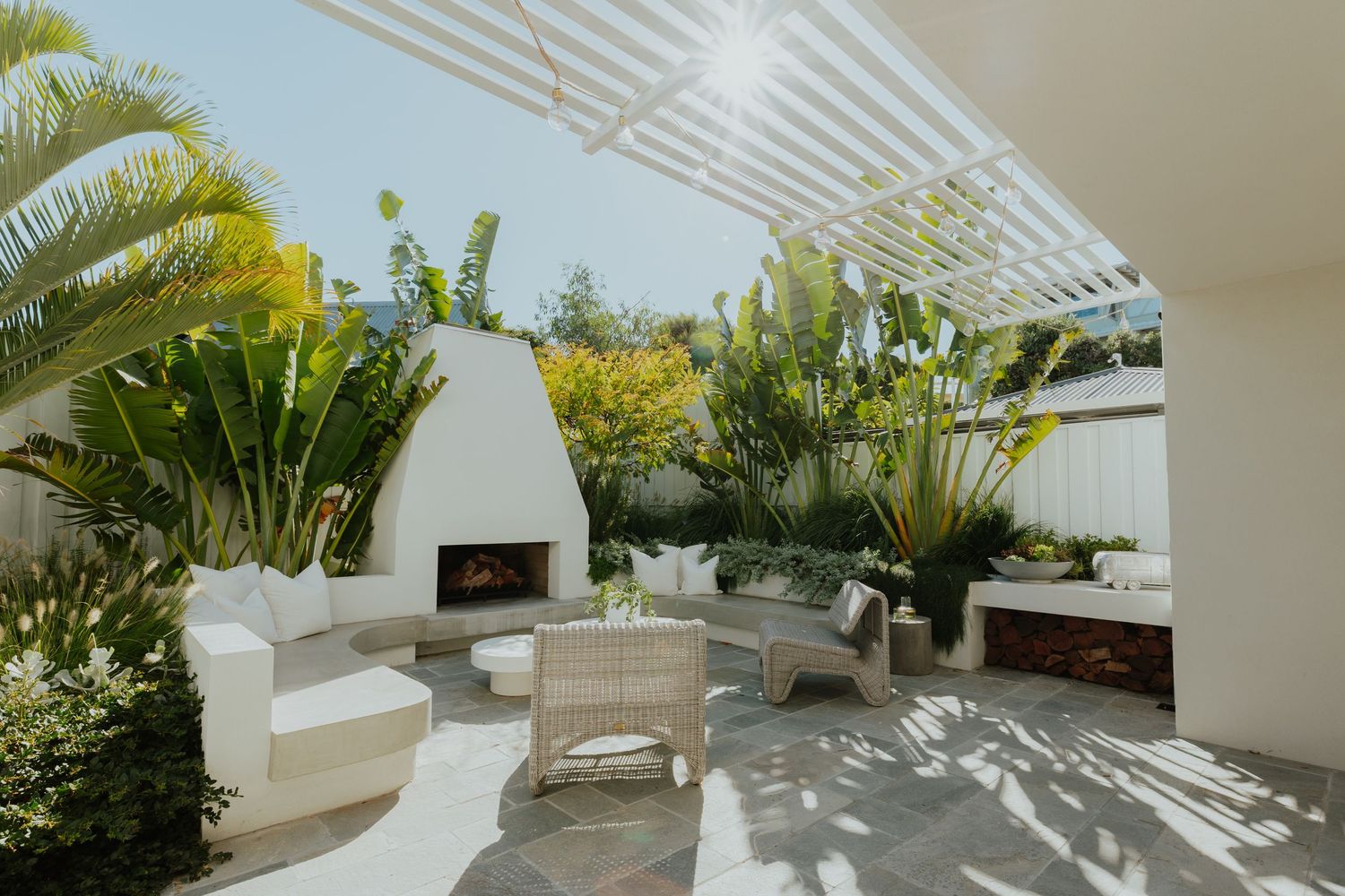 Bright outdoor patio with white pergola, wicker furniture, white fireplace, and lush tropical plants.
