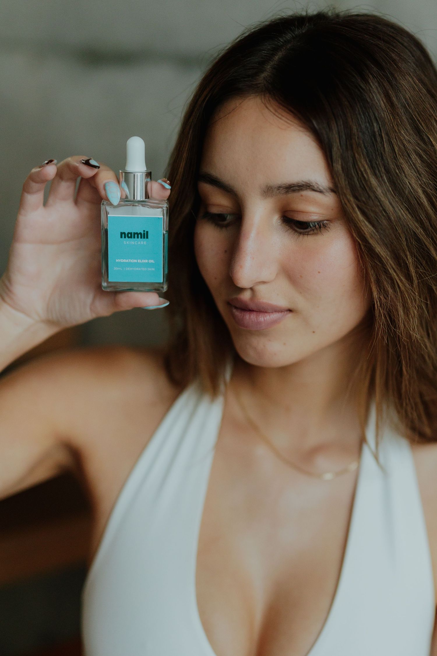 Young woman in white top holding teal Namii skincare dropper bottle close to her face.