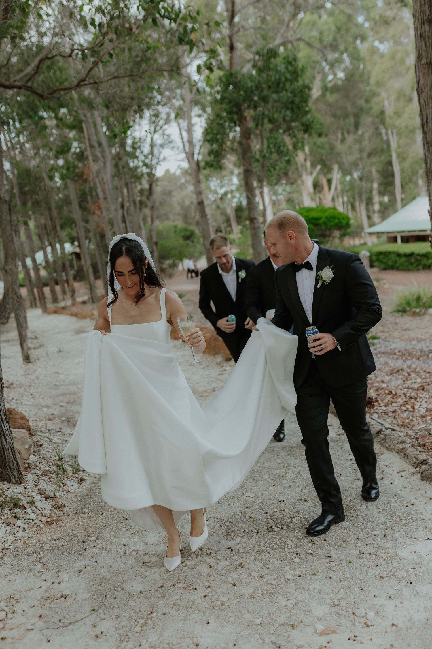 Groom carries bride's train while walking through forest path with groomsman at outdoor wedding.