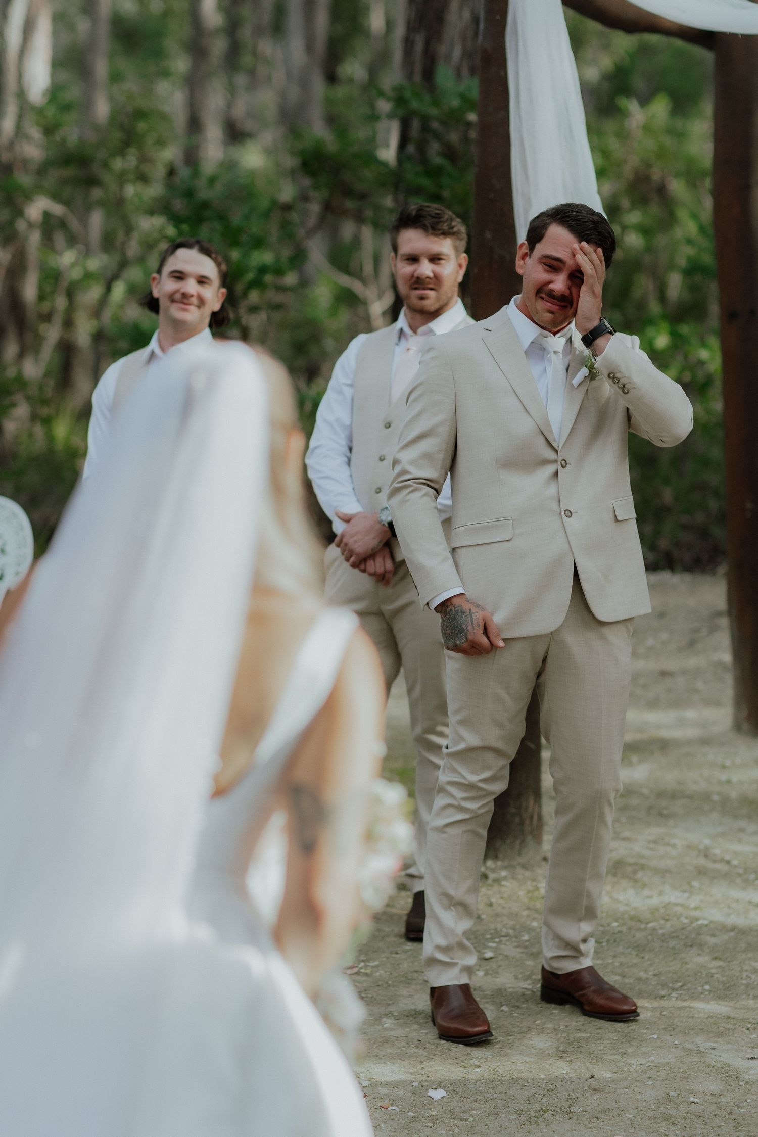 Emotional groom wiping tears seeing his bride walk down the aisle at outdoor wedding ceremony.