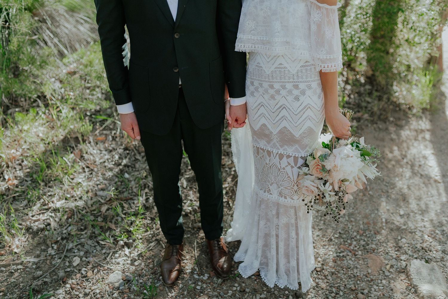 Close-up of bride in lace dress and groom holding hands outdoors.