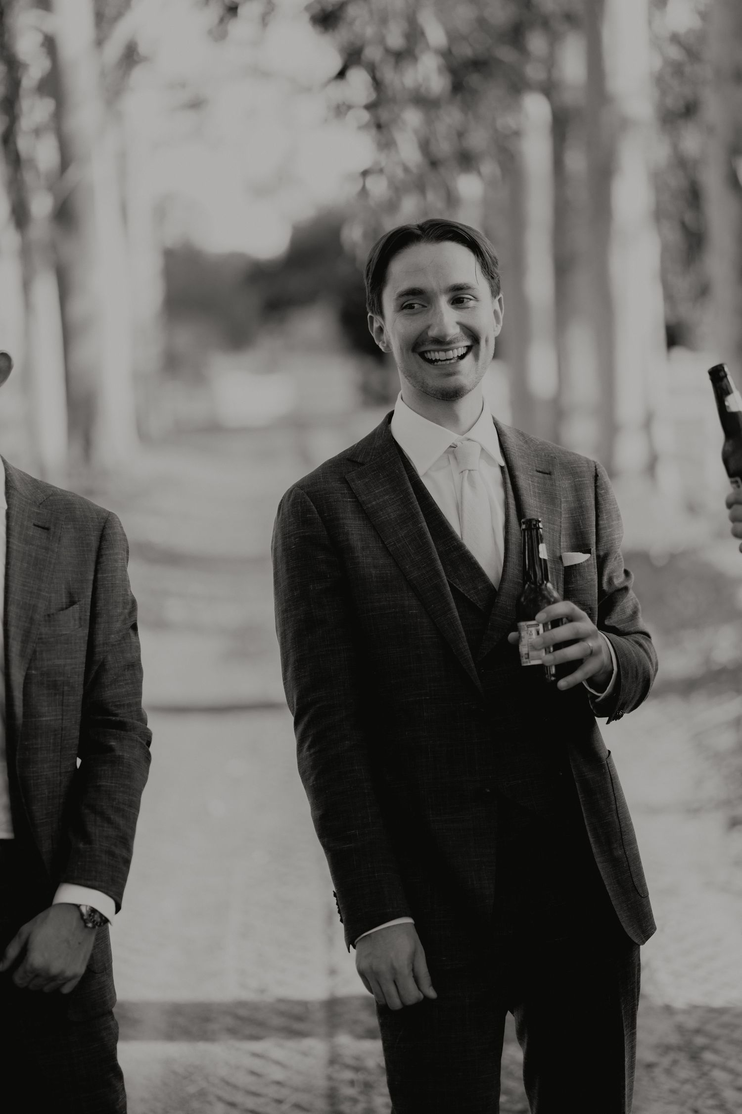 Smiling groomsman in a plaid suit holding a beer bottle at an outdoor wedding, black and white photo.