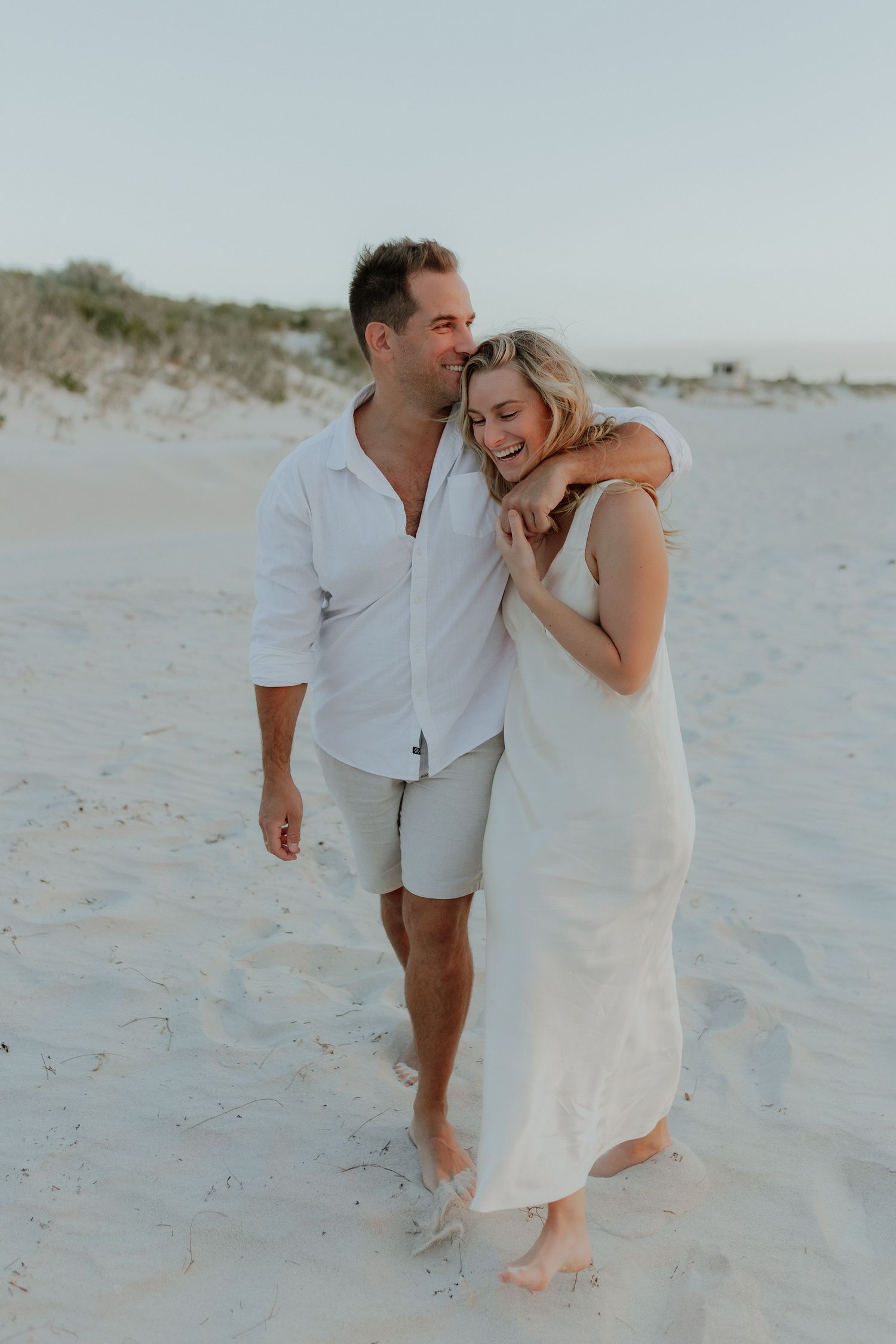 Couple laughing and walking together on a white sand beach at sunset in matching white outfits.
