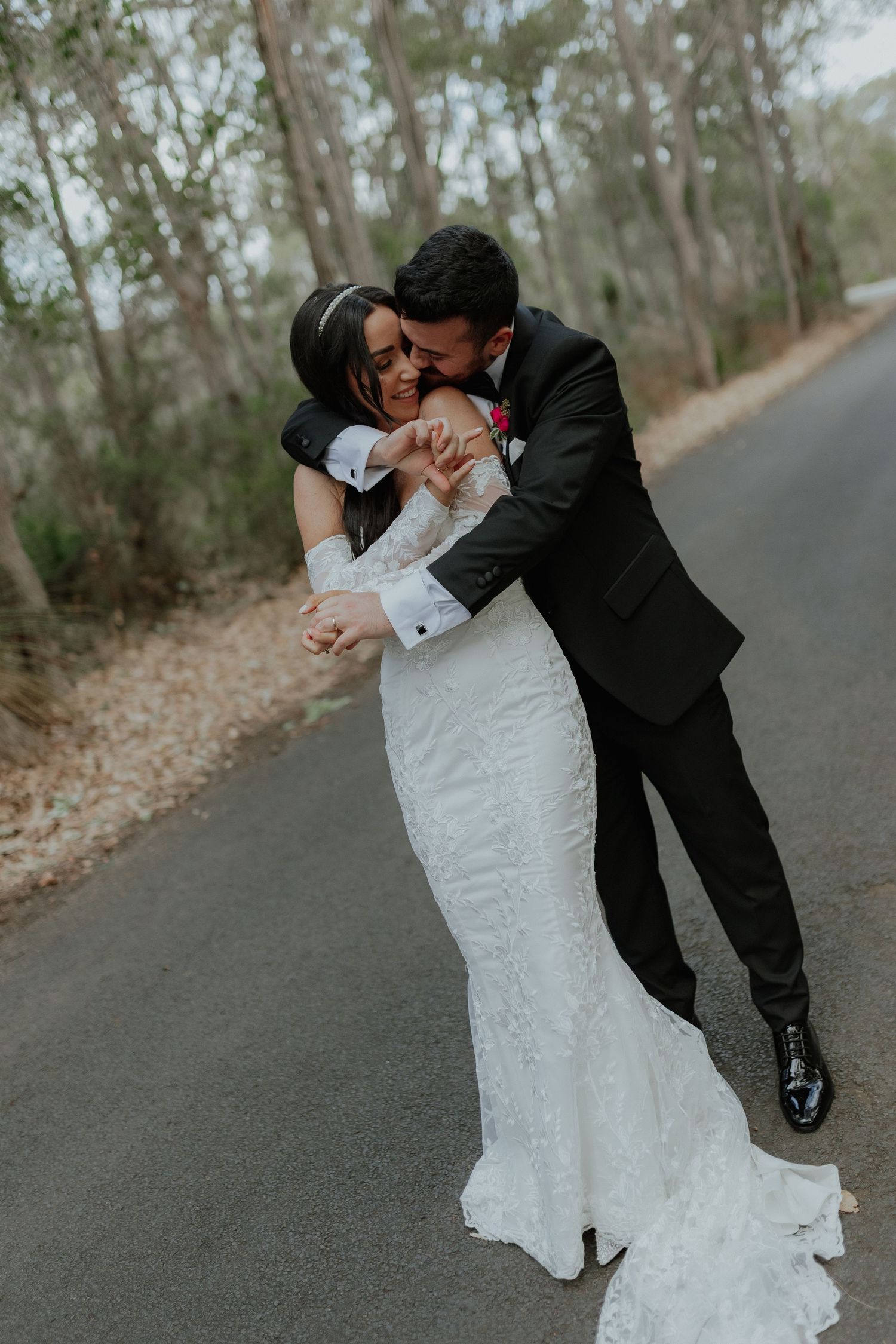 Groom in black suit dipping and embracing laughing bride in lace wedding dress on a forest road.
