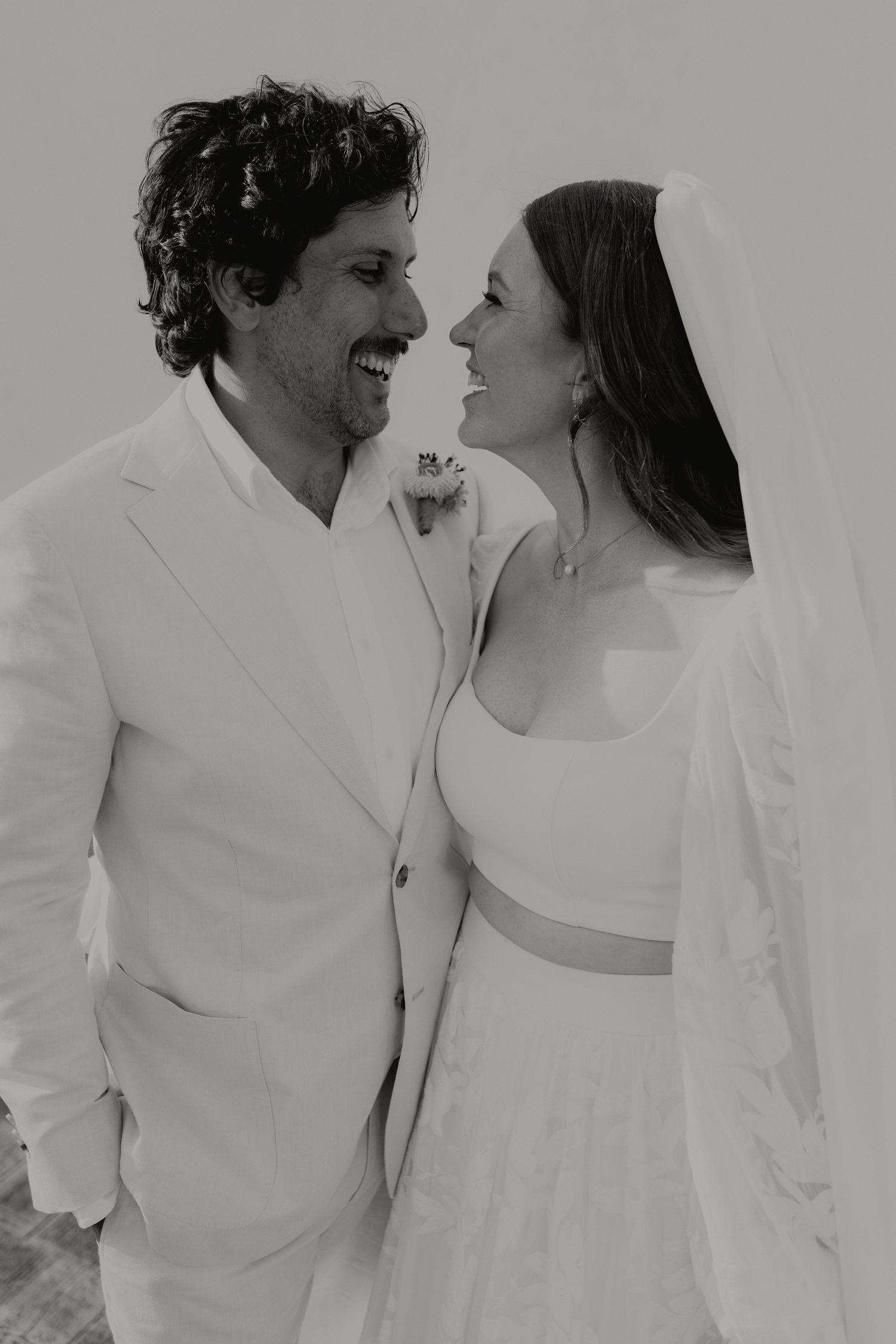Black and white photo of smiling bride and groom in white wedding attire sharing a joyful moment together.