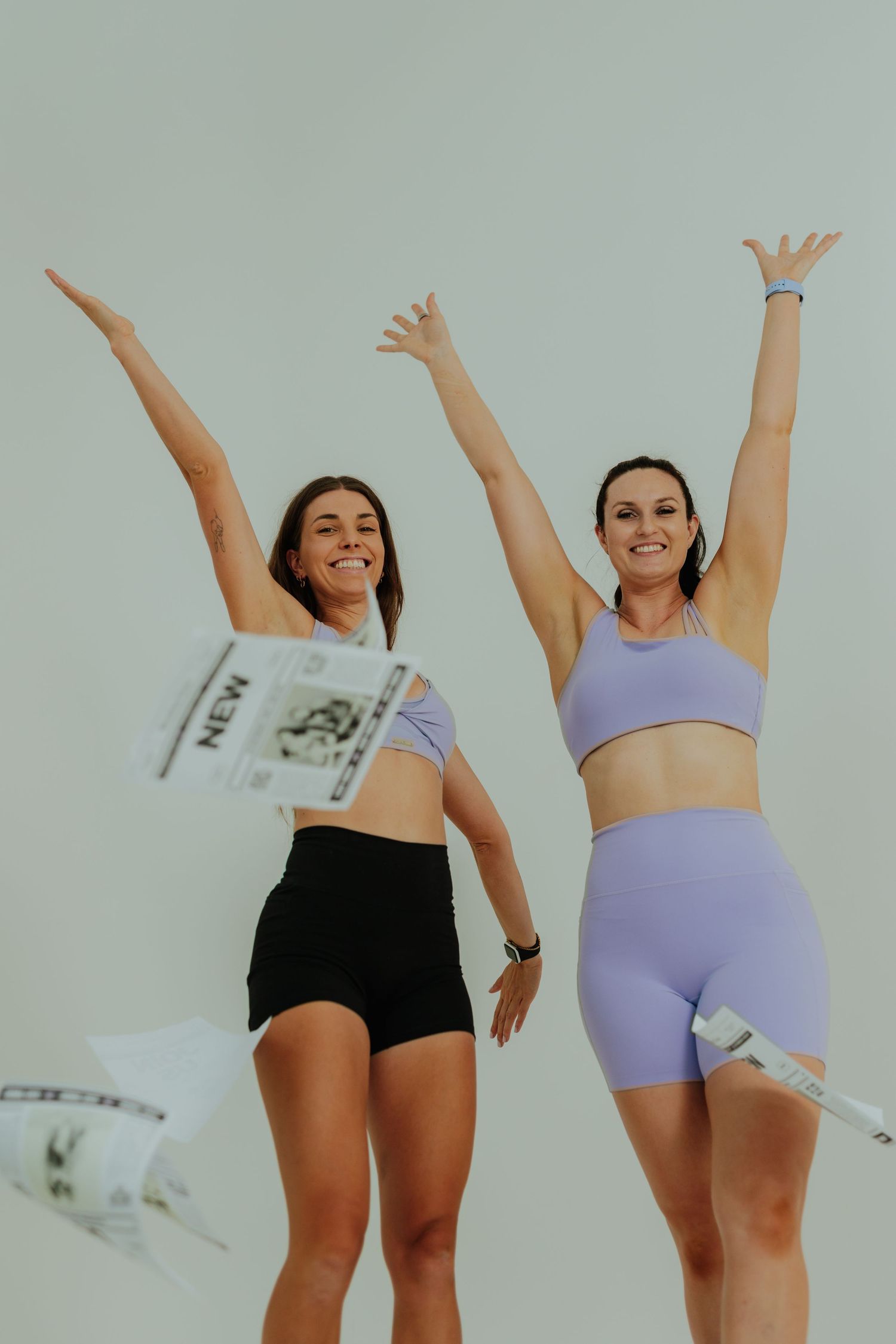 Two women in athletic wear celebrate outdoors, tossing newspapers in the air with arms raised.