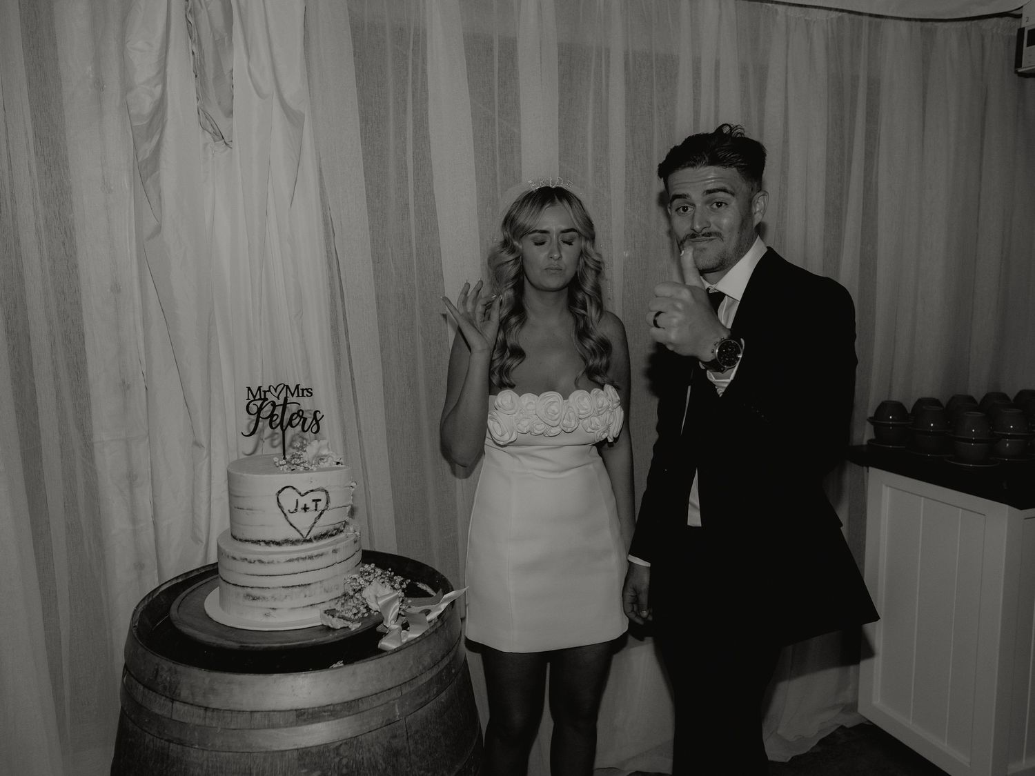 Black and white photo of bride and groom posing playfully beside tiered wedding cake on barrel.