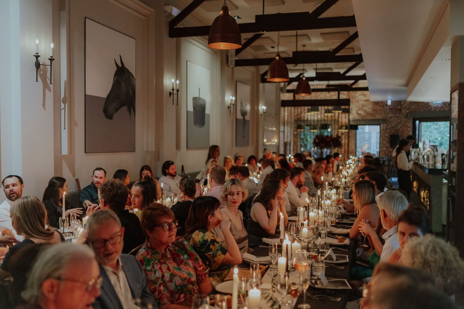 Elegant wedding reception dinner with long candlelit tables, guests dining in a warmly lit venue with horse artwork on walls.