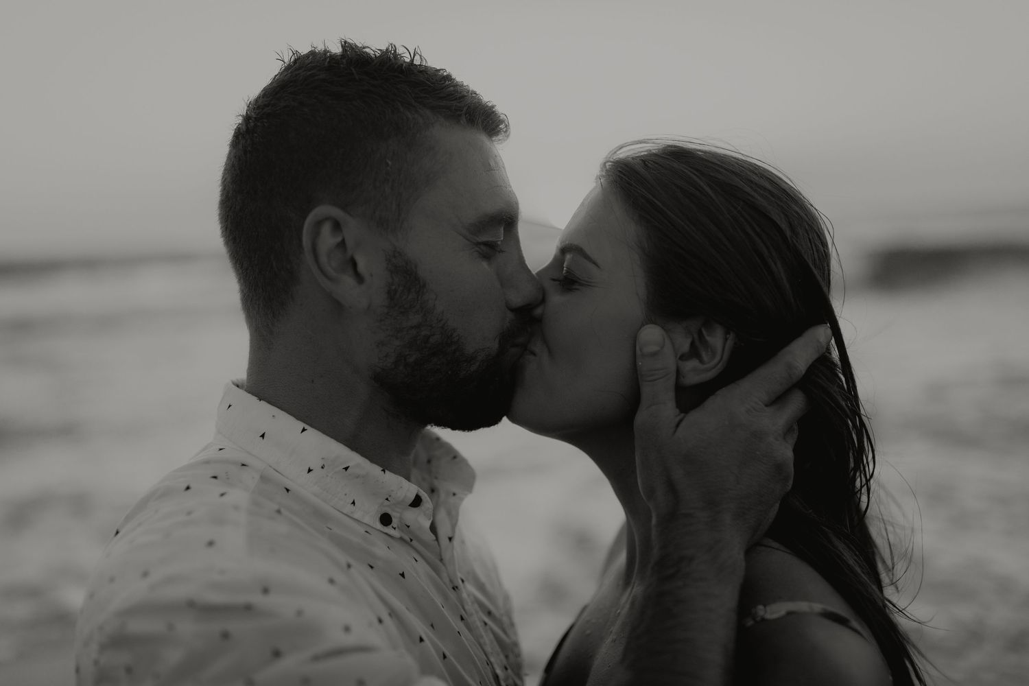 Black and white photo of a couple sharing a romantic kiss on a beach at dusk.