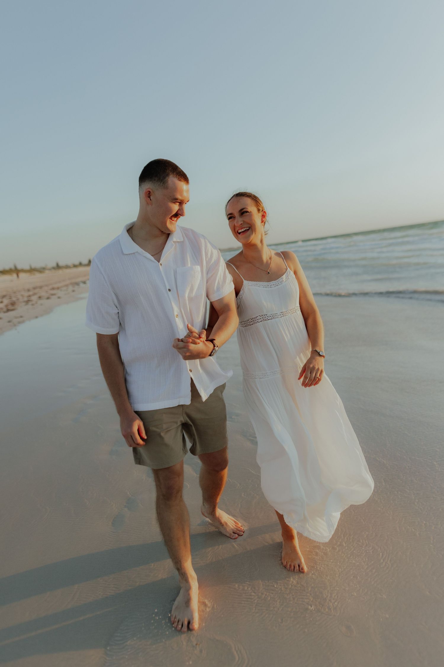 Smiling couple walking hand in hand along sandy beach at sunset, woman in flowing white dress.