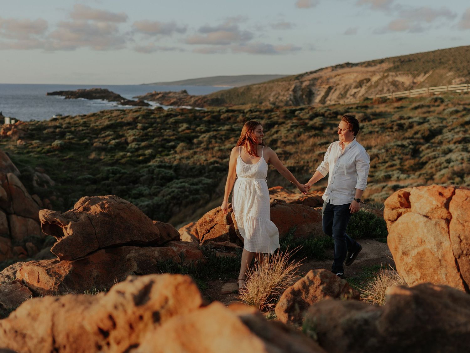 Couple holding hands on rocky coastal cliffs at golden hour with ocean views.